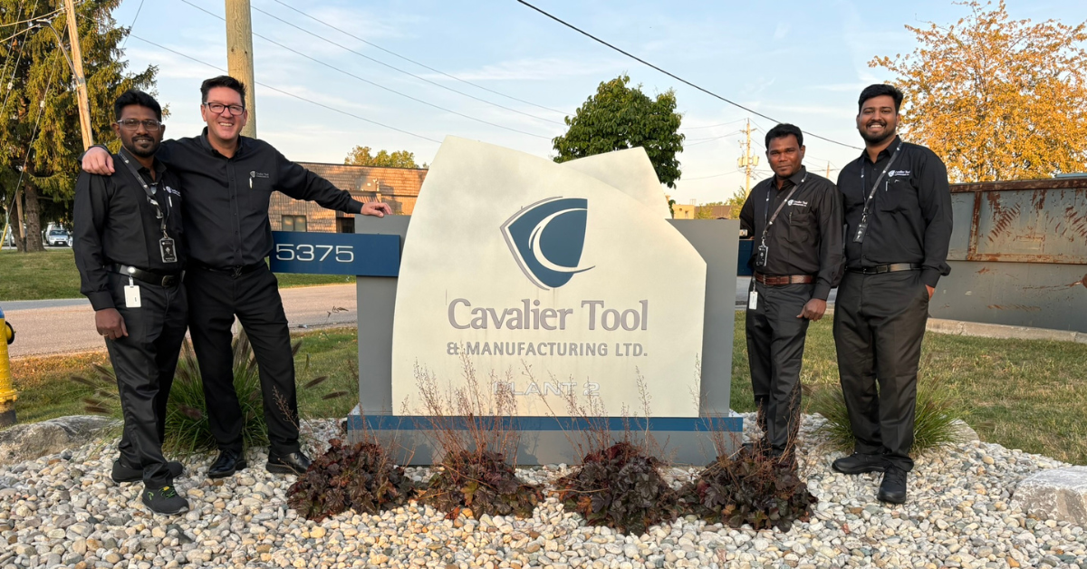 Left to Right posing in front of the sign at Cavalier Plant 2: Vella, Brian Bendig, Mani, and Nandha. They are all wearing the Cavalier Tool Uniform (A black button down shirt with a silver Cavalier shield on the left upper chest. The sign is silver and blue with the vertical Cavalier Logo in the middle. The sign is shaped like a shield.